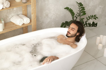 Young relaxed man with closed eyes lying in bathtub with hot water and foam