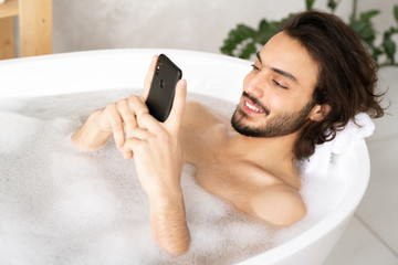 Cheerful guy with smartphone watching video or making selfie while taking bath