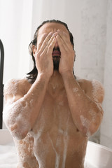 Wet young muscular man keeping his hands by face while washing his body