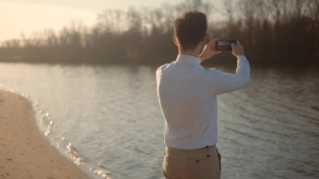 Businessman Takes Pictures On Smartphone On City Beach.Man Taking Mobile Photo.Man Holding Mobile Phone And Taking Picture At Sunset.Close Up Attractive Male Happy Using Mobile App For Photo Or Video.