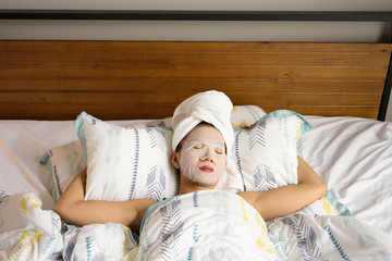 Asian woman sleeping wearing facial treatment whitening marks covered face with towel around her hair in bed room background.