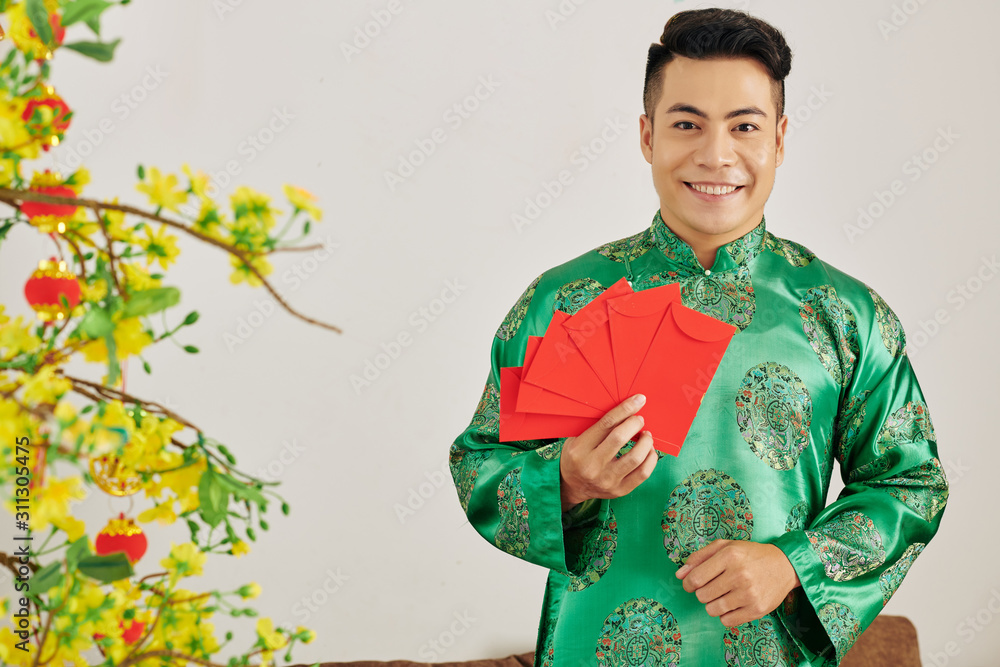 Poster Portrait of young attractive Asian man holding red envelopes for Tet celebration