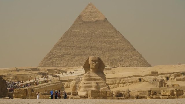 The Great Sphinx with massive pyramid of Giza in the background and crowds of tourists visiting this historical location. Hot desert winds and erosion of antiquities and structures.
