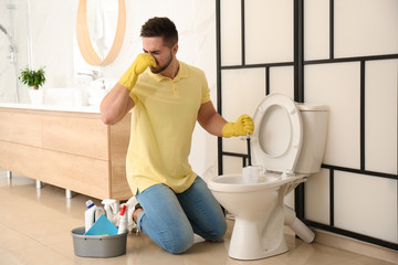 Young man feeling disgust while cleaning toilet bowl in bathroom