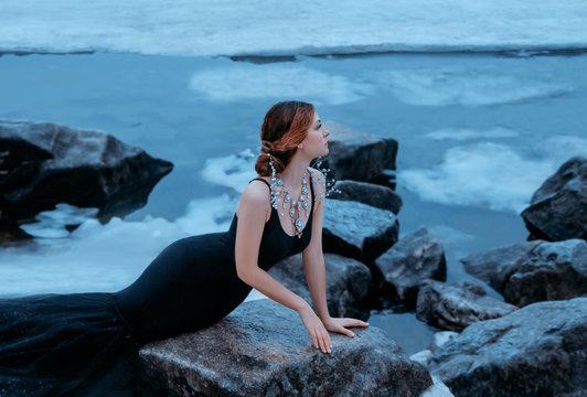 red haired mermaid girl sits on shore of frozen river waits for change. concept of love hope. Black luxurious elegant bodycon dress. Creative jewelry necklace precious stones diamond silver beads