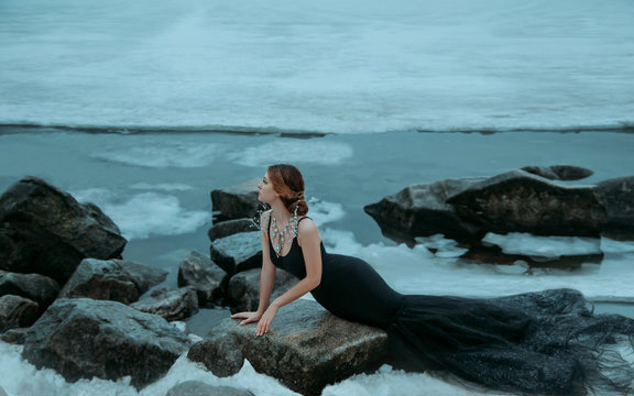 Sexy Woman long black dress silhouette mermaid sitting on stone. backdrop river ice water cold. Lady Elegant sea queen enjoying winter. Creative jewelry gemstone necklace royal luxury collar beads