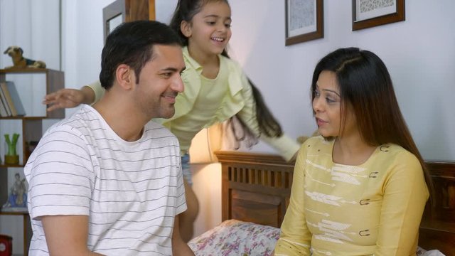 4K Stock Footage of a happy nuclear Indian family in their bedroom - Young cute daughter and young indian parents. Indian Stock Video of a girl child running to hug her parents sitting on the bed. ...