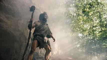 Tuinposter Primeval Caveman Wearing Animal Skin Holds Stone Hammer Stands Near Cave and Looks Around Prehistoric Landscape, Ready to Hunt Animal Prey. Neanderthal Going Hunting into Jungle. Low Angle Shot © Gorodenkoff