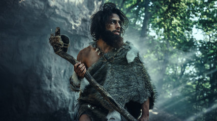 Primeval Caveman Wearing Animal Skin Holds Stone Tipped Hammer Comes out of the Cave and Looks Around Prehistoric Landscape, Ready to Hunt Animal Prey. Neanderthal Going to Hunt in the Jungle