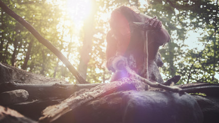 Silhouette of Primeval Caveman Wearing Animal Skin Trying to make a Fire with Bow Drill Method. Neanderthal Kindle First Man-Made fire in the Human Civilization History. Low Angle Shot