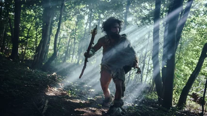 Foto op Plexiglas Primeval Caveman Wearing Animal Skin Holds Stone Tipped Spear Looks Around, Explores Prehistoric Forest in a Hunt for Animal Prey. Neanderthal Going Hunting in the Jungle © Gorodenkoff