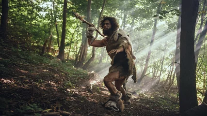 Foto op Canvas Primeval Caveman Wearing Animal Skin Holds Stone Tipped Spear Looks Around, Explores Prehistoric Forest in a Hunt for Animal Prey. Neanderthal Going Hunting in the Jungle © Gorodenkoff