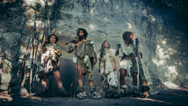Tribe of Four Hunter-Gatherers in Animal Skin Holding Stone Tipped Tools, Pose at Entrance of their Cave. Portrait of Neanderthal Family and their Way of Living. The Shot is Done with Cold Filter