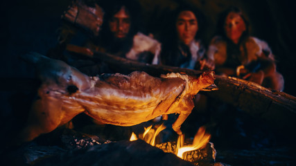 Close-up Shot of Homo Sapiens Family Cooking Animal Meat over Bonfire and then Eating it. Tribe of Prehistoric Hunter-Gatherers Wearing Animal Skins Eating in a Dark Scary Cave at Night.