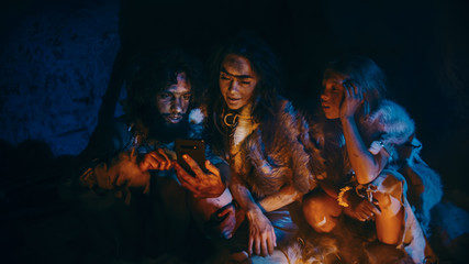 Tribe of Prehistoric, Primitive Hunter-Gatherers Wearing Animal Skins Use Smartphone in a Cave at Night. Neanderthal / Homo Sapiens Family Browsing Internet on Mobile Phone, Cooking Food over Fire