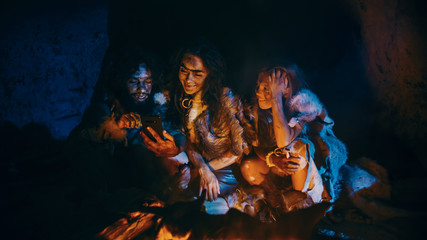 Tribe of Prehistoric, Primitive Hunter-Gatherers Wearing Animal Skins Use Smartphone in a Cave at Night. Neanderthal / Homo Sapiens Family Browsing Internet on Mobile Phone, Cooking Food over Fire