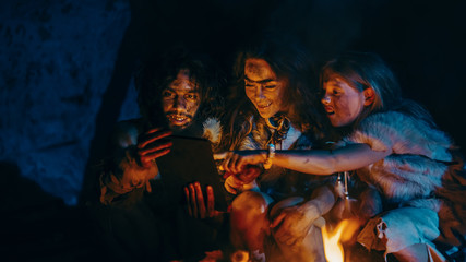 Fototapeta na wymiar Tribe of Prehistoric, Primitive Hunter-Gatherers Wearing Animal Skins Use Digital Tablet Computer in a Cave at Night. Neanderthal or Homo Sapiens Family Browsing Internet, Watching Videos, TV Shows 