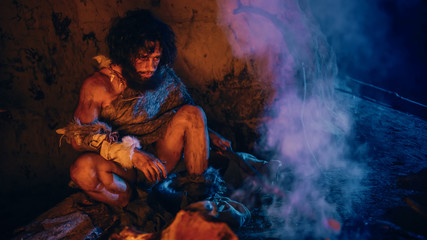 Portrait of Primeval Caveman Wearing Animal Skin Sitting in His Cave, Warms Up Near the Fire....