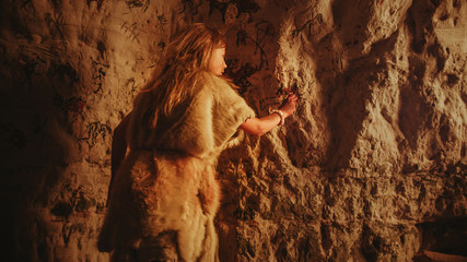 Back View of a Primitive Prehistoric Neanderthal Child in Animal Skin Draws Animals and Abstracts on the Walls at Night. Creating First Cave Art with Petroglyphs, Rock Paintings Illuminated by Fire.