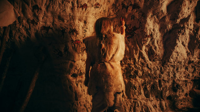 Back View of a Primitive Prehistoric Neanderthal Child Wearing Animal Skin Draws Animals and Abstracts on the Walls at Night. Creating First Cave Art with Petroglyphs, Rock Paintings.
