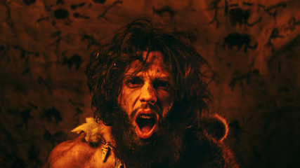 Portrait of Primeval Caveman Wearing Animal Skin Standing in His Cave At Night, Holding Torch with Fire and Attacking. Primitive Neanderthal Hunter / Homo Sapiens Screaming and Threatening