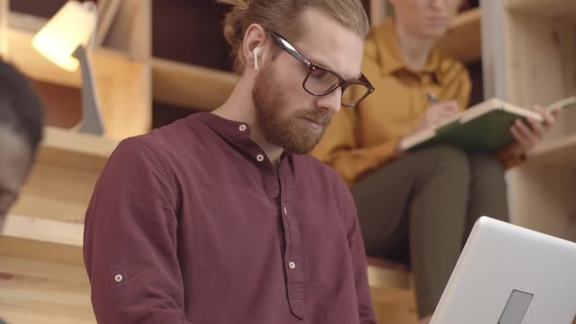 Tilting waist-up shot of diverse students sitting on benches in cozy coworking lounge &ndash; red-haired hipster guy with beard working on laptop, and thoughtful Caucasian girl writing in notebook