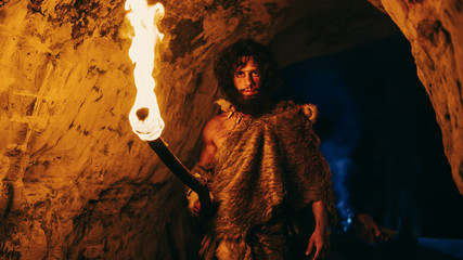Portrait of Primeval Caveman Wearing Animal Skin Exploring Cave At Night, Holding Torch with Fire...