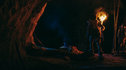 Primeval Caveman Wearing Animal Skin Stands in a Cave At Night, Holding Torch with Fire Looking Out...