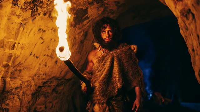 Portrait of Primeval Caveman Wearing Animal Skin Exploring Cave At Night, Holding Torch with Fire Looking into Camera at Night.