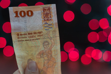 Hand holds a 100 Sri Lanka rupees bank note currency money, pink and red bokeh background with copyspace