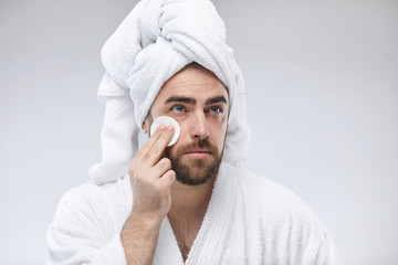 Young man wearing white bathrobe with towel headwrap swiping cotton pad gently over his face looking away studio portrait shot © Seventyfour