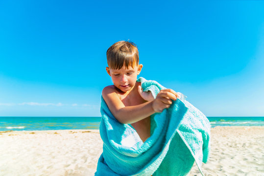 The Boy On The Beach Hides With A Towel And Wipes Himself From Sea Water After Swimming In The Sea.