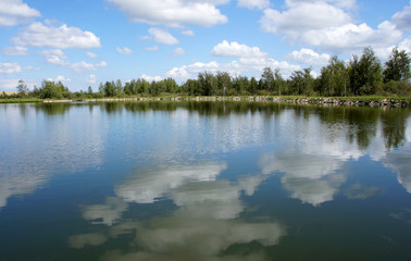 The green shore of the lake against the blue sky with white fluffy clouds is reflected in the clear water. Beautiful scenery.