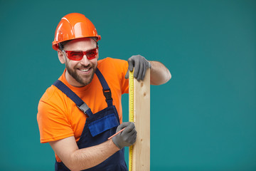 Horizontal studio portrait of professional carpenter sizing wooden plank with measuring tape looking at camera smiling