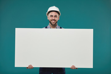 Horizontal studio portrait shot of happy young adult manual worker holding blank poster looking at...