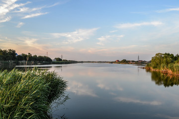 A large river with green reeds on the shore. A blue sky with light clouds reflected in the water.