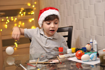 Little cute kid decorating Christmas balls at home kitchen on the yellow lights background