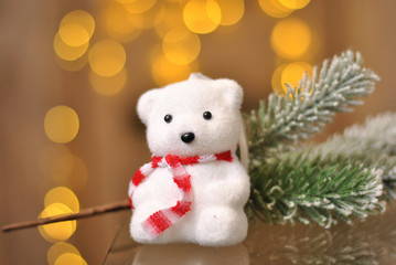 Little cute white bear wearing red scarf  toy with snowed fir tree branch on the yellow garland lights background. Christmas holiday and New Year decoration