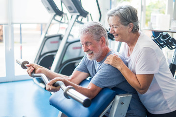 Obraz premium beautiful couple of two seniors at the gym doing exercises with a strong light at the background - woman hugging her husband holding a barr - active lifestyle