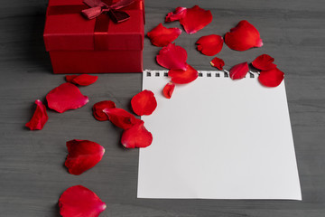 Empty sheet of paper for text and red gift box next to a bouquet and red rose petals against a dark background.