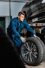 selective focus of young mechanic with car wheel near raised car in workshop