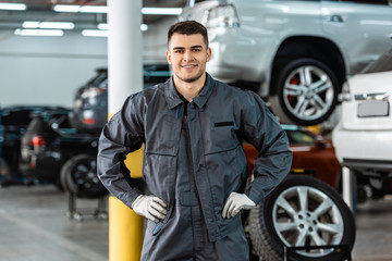 smiling mechanic looking at camera while standing in workshop with hands on hips