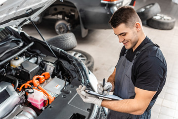 smiling mechanic writing on clipboard while inspecting car engine compartment