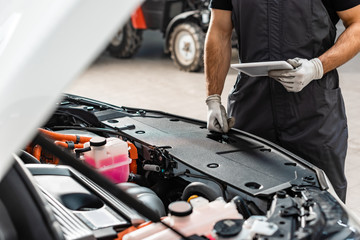 partial view of mechanic holding digital tablet near car engine compartment