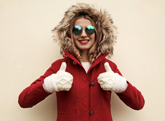 Portrait happy smiling young woman showing thumbs up wearing red jacket with fur hood and white mittens over background