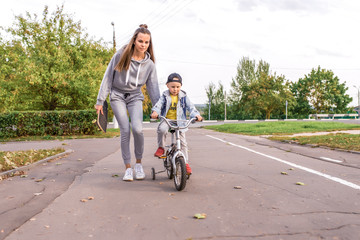 Young woman mom teaches child ride bicycle, little boy son. Rest on weekend in fall in park. Everyday warm clothes, autumn day. Support parental care and upbringing.