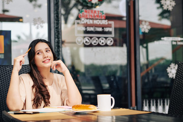 Pensive young smiling female student doing homework when sitting at cafe table with cup of coffee