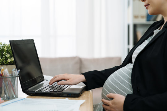 Young asian pregnant business woman using laptop computer at modern office. focus on big pregnancy belly with one hand holding sitting at work desk indoors. close up maternity abdomen by notebook pc