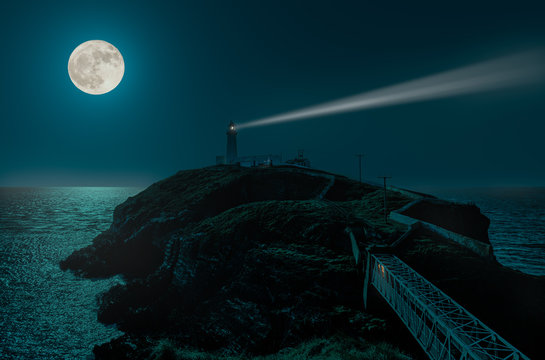 A full moon night at the South Stack lighthouse on the Irish Sea. The moonlight is reflected by the sea. The light beam can be seen. At the end of the bridge there is light in the tunnel.