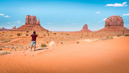 Amazed young Latino American traveler in flip flops takes photo of famous Monument Valley desert landscape of large red rock domes on Utah-Arizona border, United States of America